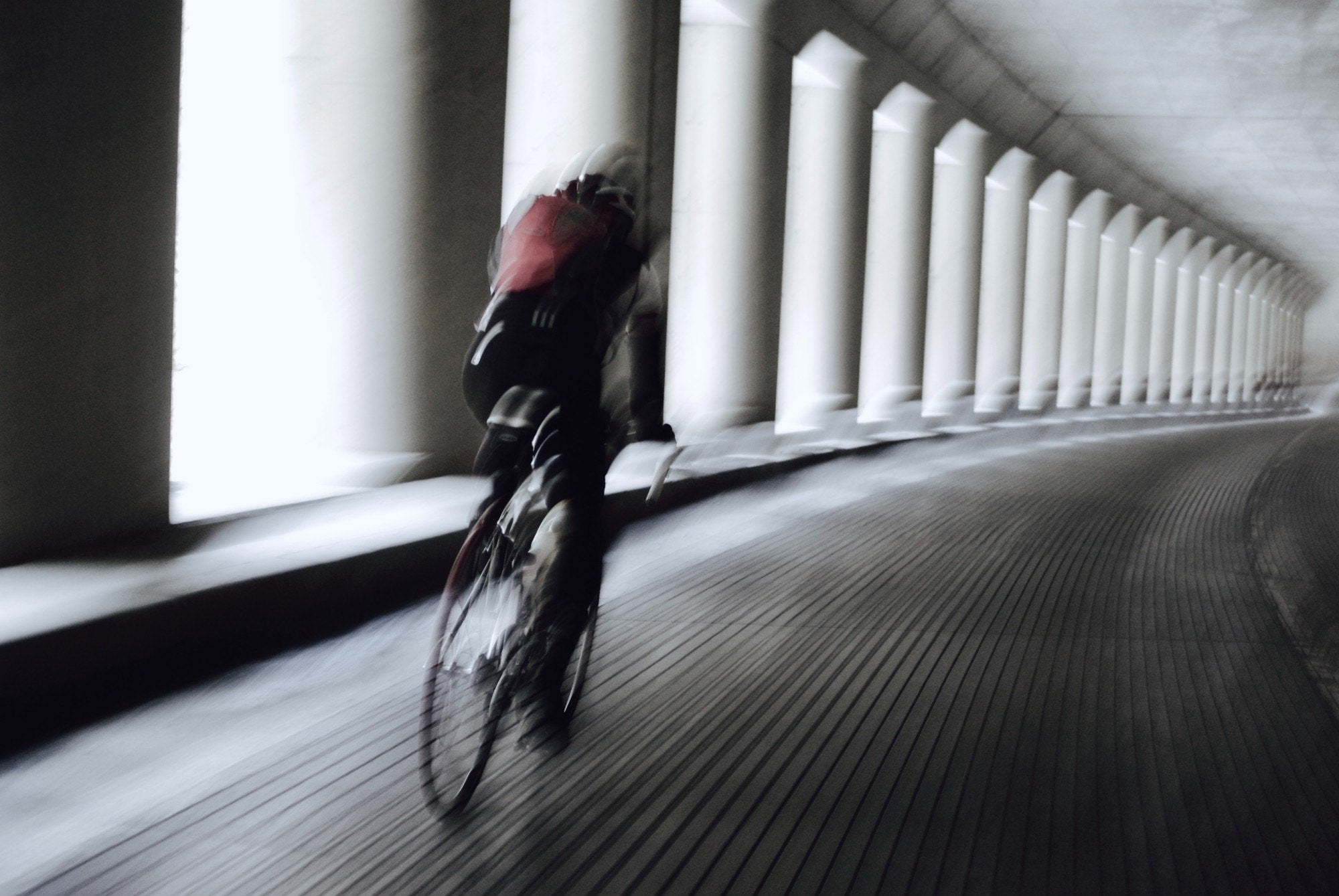 A cyclist in motion through a long colonnade, black and white with a red backpack